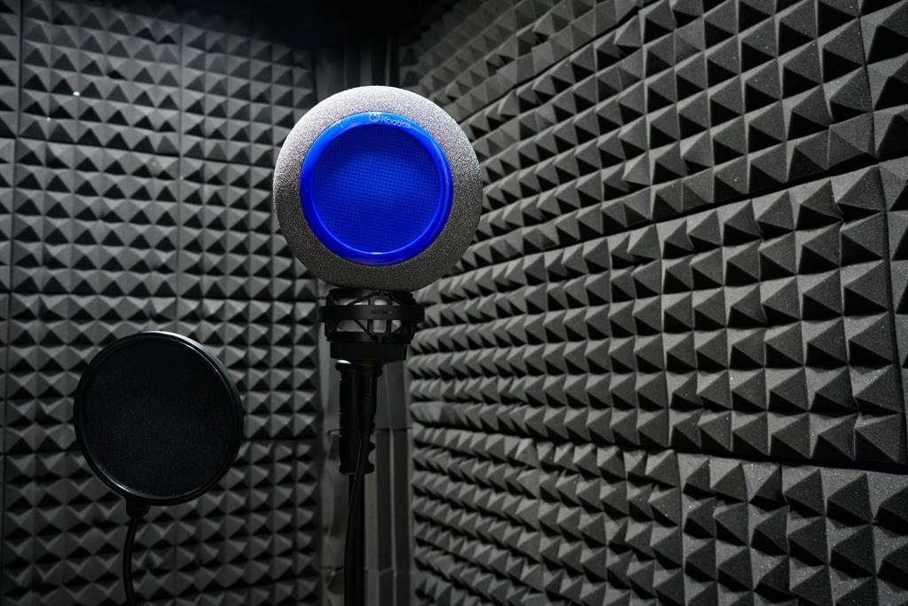 The image displays a close-up of a professional recording studio's microphone setup. There's a high-quality studio microphone with a blue LED-lit pop filter attached, positioned on a microphone stand. To the left is a black pop shield on its own stand, used to reduce plosive sounds during vocal recordings. The background shows an acoustic foam panel wall, in a classic wedged design, which helps to dampen and diffuse sound for optimal recording quality. The setup is indicative of a professional environment geared towards capturing clear and precise audio recordings.