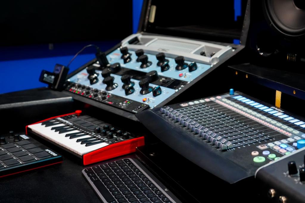 The image shows a close-up of a modern music production setup. There's a small MIDI keyboard with red accents on the side, a digital audio mixing console with illuminated buttons and sliders, and two audio interface units with various knobs and toggle switches. A pair of studio monitor speakers can be partially seen in the background, set against a blue wall. In the foreground, there's also a laptop keyboard, suggesting the integration of digital audio workstation software in the setup for music recording and editing. The equipment appears to be high-quality, indicating a professional or semi-professional studio environment.