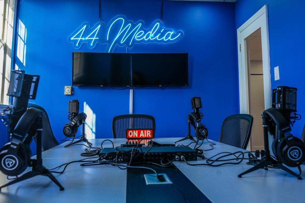 The image showcases a modern podcasting or broadcasting studio setup. A neon sign spelling out "44 Media" glows prominently against a bright blue wall. Below the sign, two black monitors are mounted on the wall. A large white table is set with multiple podcasting stations, each equipped with a professional microphone, headphones, and a pop filter. An "ON AIR" illuminated sign sits centrally on the table, indicating a live session is in progress or ready to start. The room is well-lit and offers a clean, professional environment, designed for audio recording and live broadcasting. The door on the right suggests a private, sound-controlled space, emphasizing the studio's focus on quality production.