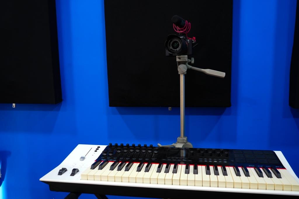 The image features a music studio setup against a vivid blue wall with two black acoustic panels. In the center, there is a white MIDI keyboard controller with black and white keys, several control knobs, and faders. To the right, mounted on a gray tripod stand, is a camera with a red shock mount microphone attached on top. The tripod has a crank handle, indicating the ability to adjust the height. The setup suggests a focus on both music production and video content creation, likely for tutorials or performances.