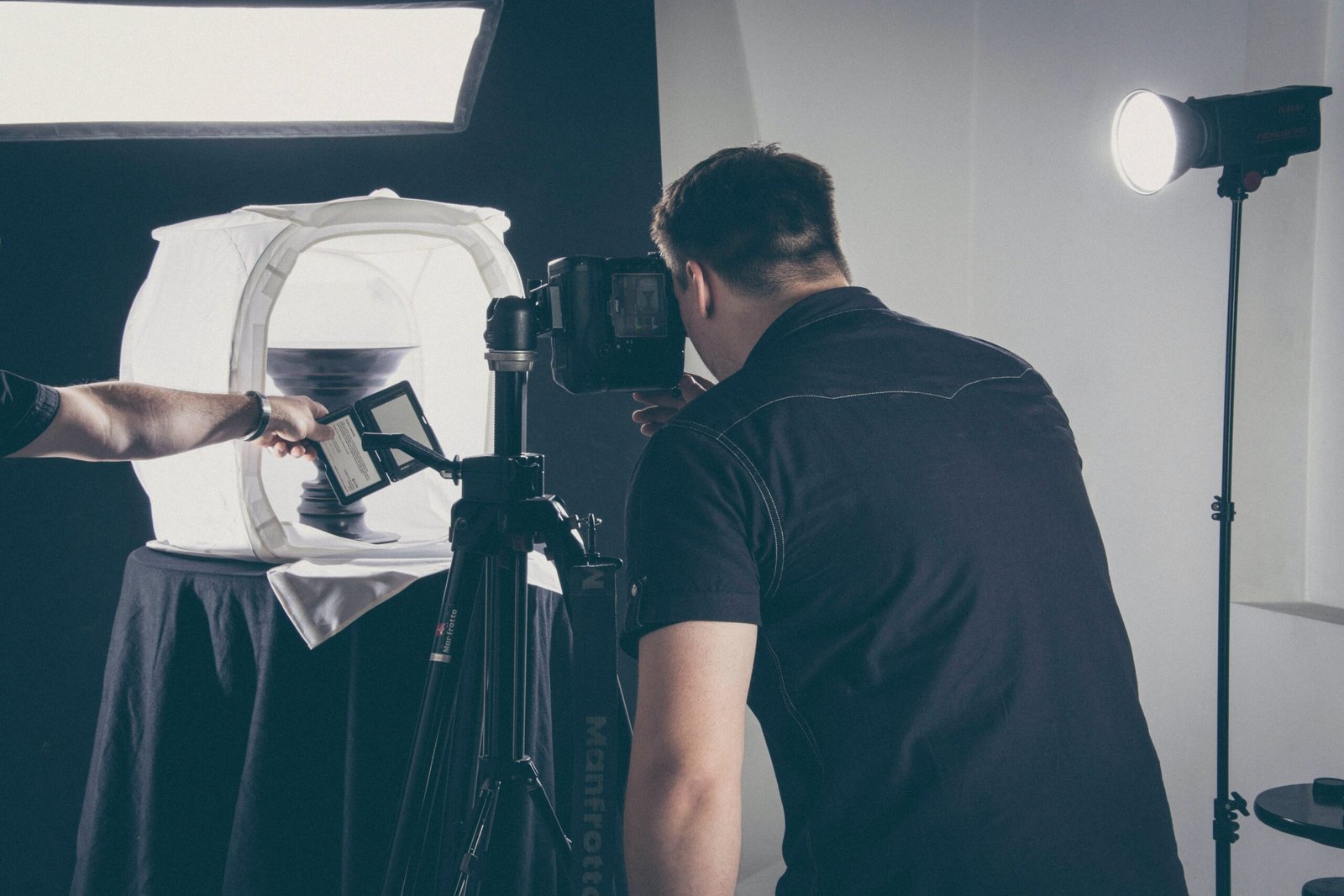 The image captures a behind-the-scenes moment of a product photography shoot. A photographer is focused on composing a shot on a digital camera mounted on a tripod. In the foreground, a hand is holding a tablet with a reference image, suggesting a comparison or duplication of a specific shot. The product, obscured by a light diffusing tent to ensure even illumination, is placed on a pedestal against a dark backdrop, ensuring focus on the item. Off to the side, a studio light on a stand provides additional lighting, contributing to the professional setup. The overall mood suggests meticulous attention to detail to achieve the perfect product image.